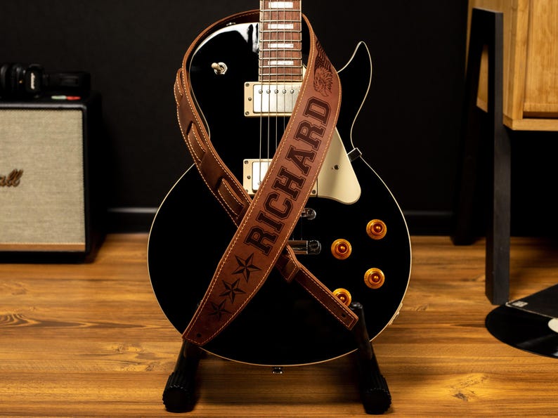 K&ouml;nnte beinhalten: Eine schwarze E-Gitarre mit einem braunen Lederriemen. Der Riemen ist mit dem Namen "RICHARD" und Sternmotiven gepr&auml;gt. Die Gitarre hat goldfarbene Kn&ouml;pfe und steht auf einem schwarzen St&auml;nder.