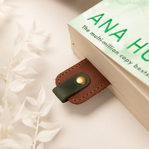 May include: A brown leather bookmark with a dark green loop and a gold-colored button is inserted into a book. The book's title is "ANA HUANG" in green letters. White foliage is in the background.