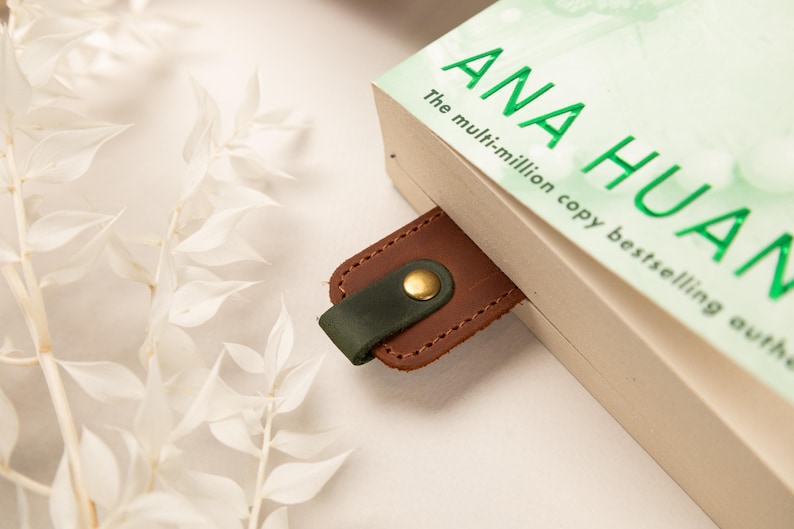 May include: A brown leather bookmark with a dark green loop and a gold-colored button is inserted into a book. The book's title is "ANA HUANG" in green letters. White foliage is in the background.