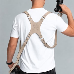 Könnte beinhalten: Ein beigefarbener Kameragurt aus Leder mit Kreuzdesign auf dem Rücken, ausgestattet mit Metallnieten und Schnallen. Das mittlere Rückenteil ist mit dem Text "FOCUS CREATE REPEAT" geprägt. Der Gurt verfügt über verstellbare Riemen für zwei Kameras.