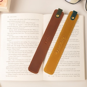 May include: Two leather bookmarks rest on an open book. One is brown with "Read" embossed, the other is mustard yellow with "One more." The bookmarks have a dark green accent and gold rivet. Glasses and a book are in the background.