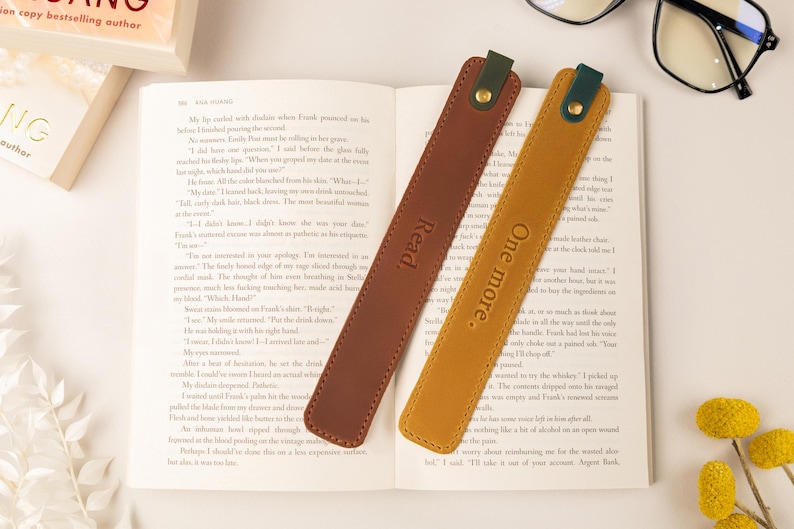 May include: Two leather bookmarks rest on an open book. One is brown with "Read" embossed, the other is mustard yellow with "One more." The bookmarks have a dark green accent and gold rivet. Glasses and a book are in the background.