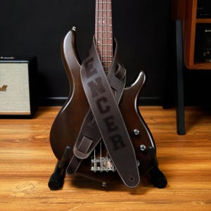 May include: A brown bass guitar with a matching leather strap. The strap is embossed with the name "Spencer" in large, white-stitched letters. The guitar is on a black stand, with a wooden floor in the foreground.