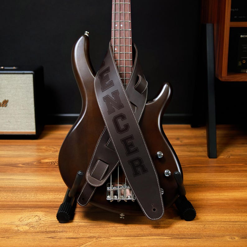 May include: A brown bass guitar with a matching leather strap. The strap is embossed with the name "Spencer" in large, white-stitched letters. The guitar is on a black stand, with a wooden floor in the foreground.