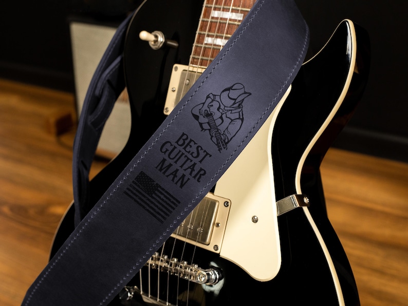 May include: A navy blue guitar strap with the text "BEST GUITAR MAN" and an illustration of a cowboy playing a guitar. The strap also features an American flag design. The strap is draped over a black electric guitar.