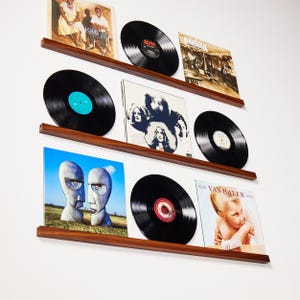 May include: A display of vinyl records on wooden shelves. The records are black with colorful labels and album art. The shelves are a dark wood color and mounted on a white wall. The album covers feature various bands and artists.