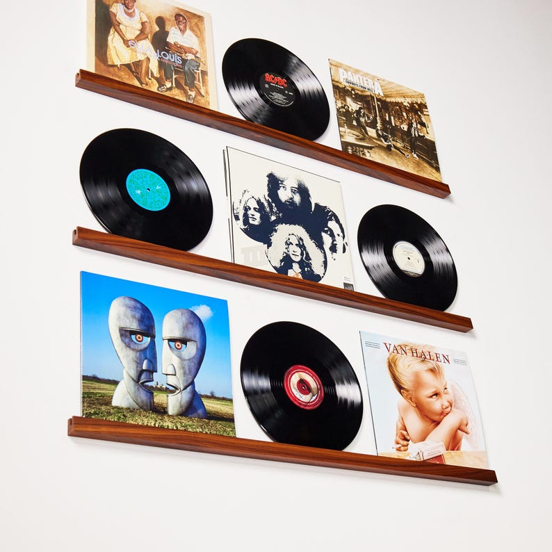 May include: A display of vinyl records on wooden shelves. The records are black with colorful labels and album art. The shelves are a dark wood color and mounted on a white wall. The album covers feature various bands and artists.
