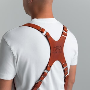 May include: A brown leather camera harness with silver metal hardware. The harness has a cross-back design with adjustable straps and a central panel with a camera graphic and the text "YOUR LOGO".