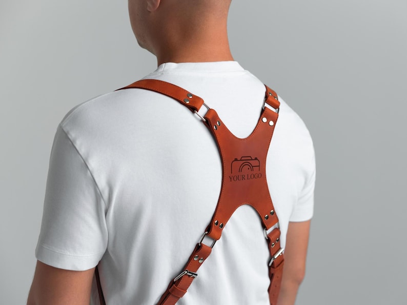 May include: A brown leather camera harness with silver metal hardware. The harness has a cross-back design with adjustable straps and a central panel with a camera graphic and the text "YOUR LOGO".