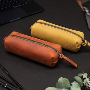 May include: Two leather pencil cases, one in a warm brown and the other in a mustard yellow, are displayed on a black surface. Each case has a zipper closure and a small leather pull tab. The brown case is labeled "RICHARD", and the yellow case is labeled "SHARON".