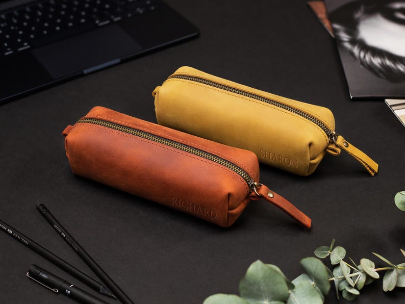 May include: Two leather pencil cases, one in a warm brown and the other in a mustard yellow, are displayed on a black surface. Each case has a zipper closure and a small leather pull tab. The brown case is labeled "RICHARD", and the yellow case is labeled "SHARON".
