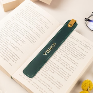May include: A dark green leather bookmark with the name "SOPHIA" in gold lettering, resting on an open book. The bookmark has a tan leather tab and a gold button. Reading glasses and yellow flowers are also in the image.