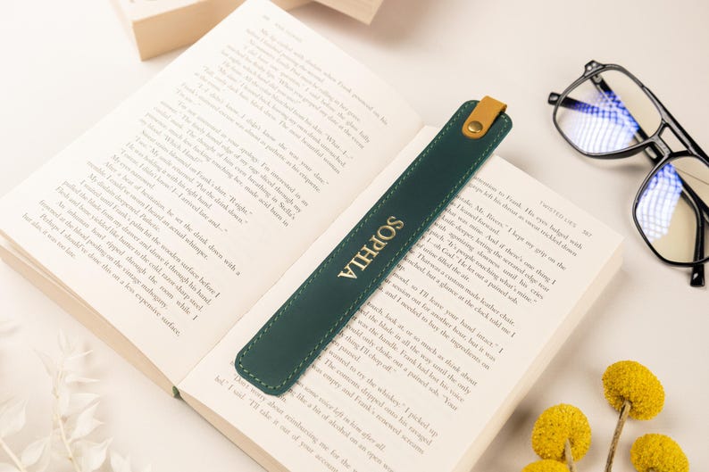 May include: A dark green leather bookmark with the name "SOPHIA" in gold lettering, resting on an open book. The bookmark has a tan leather tab and a gold button. Reading glasses and yellow flowers are also in the image.