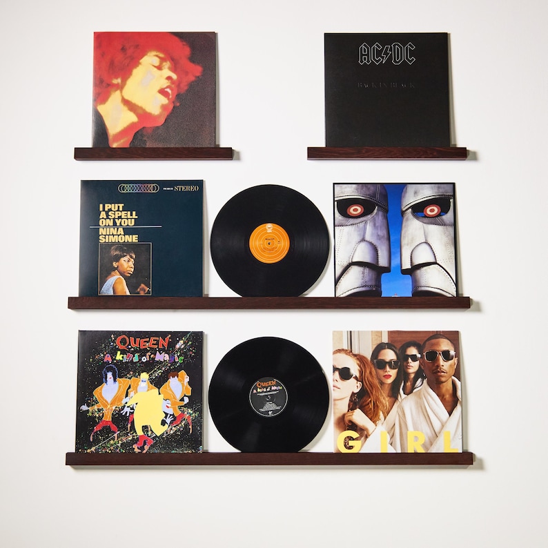 May include: A collection of vinyl records displayed on dark brown shelves against a white wall. The records feature album art from artists like Jimi Hendrix, AC/DC, Nina Simone, and Queen. The records are black with orange and yellow labels.