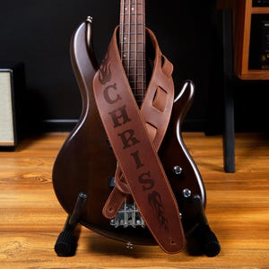 May include: A brown bass guitar with a personalized leather strap. The strap is embossed with the name "CHRIS" and a flame design. The guitar is on a wooden surface.