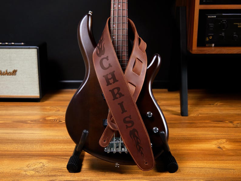 May include: A brown bass guitar with a personalized leather strap. The strap is embossed with the name "CHRIS" and a flame design. The guitar is on a wooden surface.