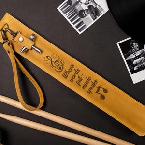 May include: Mustard yellow leather drumstick case with the quote "Where words fail - music speaks" and musical notes. Includes a wrist strap and metal hardware. Two wooden drumsticks are also visible.