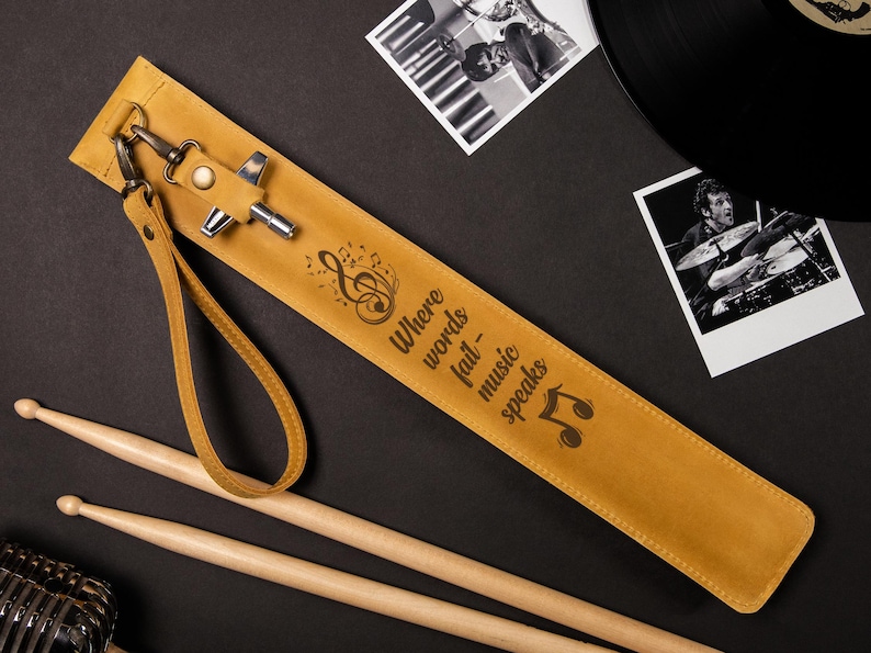 May include: Mustard yellow leather drumstick case with the quote "Where words fail - music speaks" and musical notes. Includes a wrist strap and metal hardware. Two wooden drumsticks are also visible.