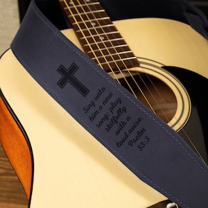 Puede incluir: Una correa de guitarra azul marino con un dise&ntilde;o de cruz y el texto "Cantadle c&aacute;ntico nuevo; ta&ntilde;ed con arte, con j&uacute;bilo." La correa est&aacute; sobre una guitarra ac&uacute;stica.