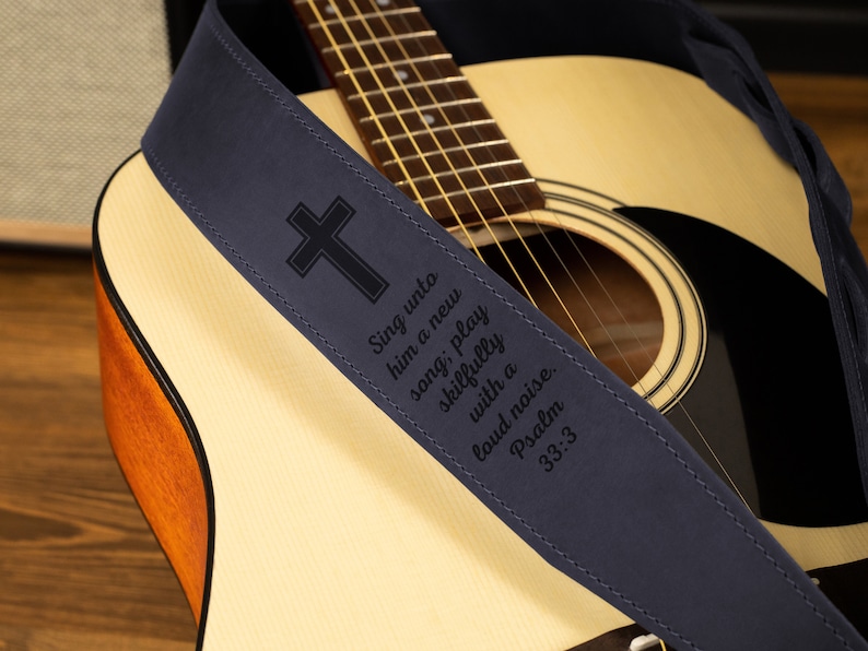 Puede incluir: Una correa de guitarra azul marino con un dise&ntilde;o de cruz y el texto "Cantadle c&aacute;ntico nuevo; ta&ntilde;ed con arte, con j&uacute;bilo." La correa est&aacute; sobre una guitarra ac&uacute;stica.