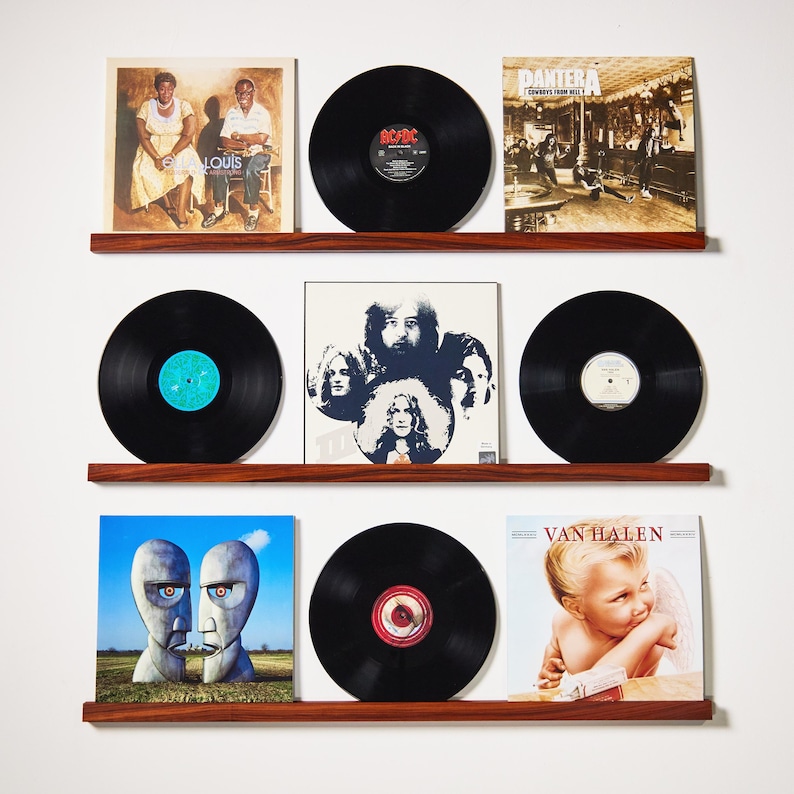 May include: A collection of vinyl records displayed on three wooden shelves. The album covers feature various artists and album art, including AC/DC, Van Halen, and Pink Floyd. The records are black with visible labels.