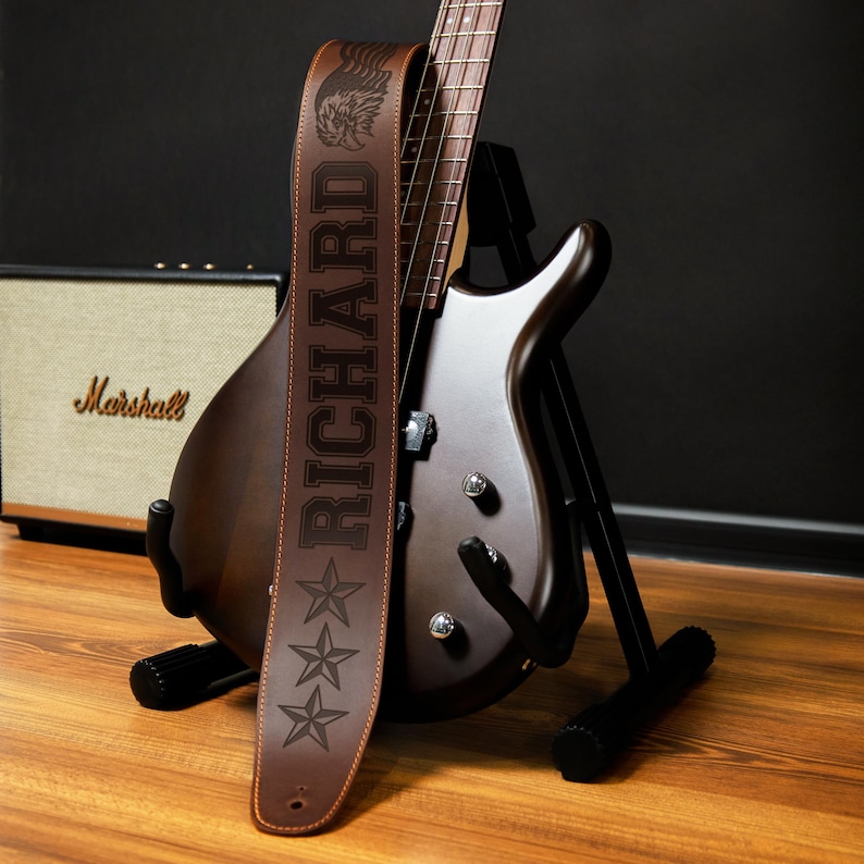 May include: A brown electric guitar with a matching leather guitar strap. The strap is embossed with the name "RICHARD" and three stars. A Marshall amplifier is in the background.