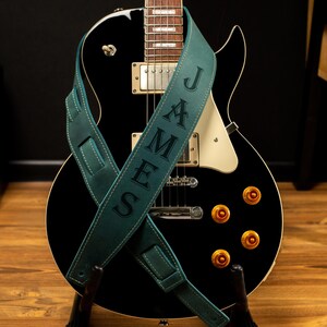 May include: A black electric guitar with gold hardware and a teal leather guitar strap. The strap is personalized with the name "JAMES" in capital letters. The guitar is on a wooden stand.