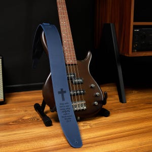 May include: A navy blue bass guitar strap with a cross and text from Psalm 33:3. The strap is draped over a brown bass guitar on a black stand. The guitar has a wooden body and neck.