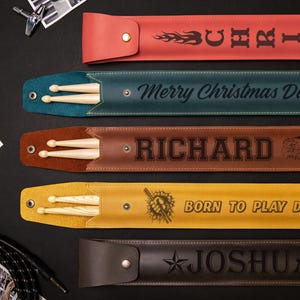 May include: A collection of personalized drumstick cases in various colors, including red, teal, brown, yellow, and dark brown. Each case features a name or phrase, such as "Chris," "Richard," and "Born to Play Drums," along with drumsticks.