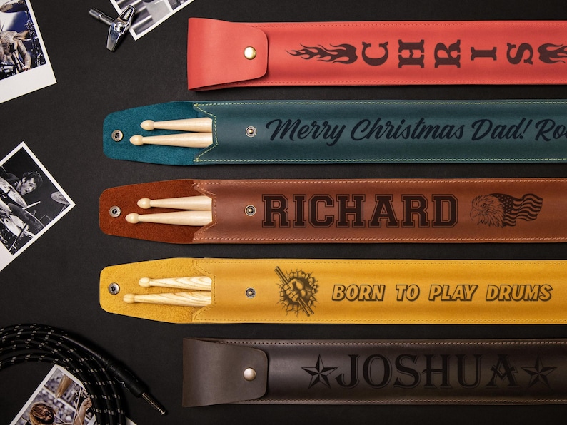 May include: A collection of personalized drumstick cases in various colors, including red, teal, brown, yellow, and dark brown. Each case features a name or phrase, such as "Chris," "Richard," and "Born to Play Drums," along with drumsticks.