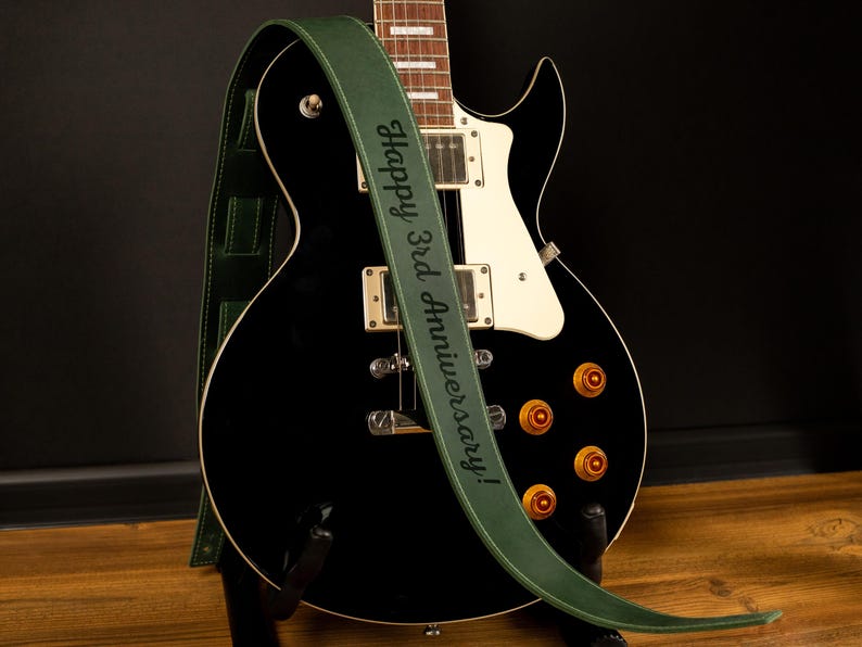 Puede incluir: Una guitarra el&eacute;ctrica negra con una correa de cuero verde. La correa tiene la inscripci&oacute;n "Happy 3rd Anniversary!" en letras blancas. La guitarra tiene perillas doradas y detalles blancos.