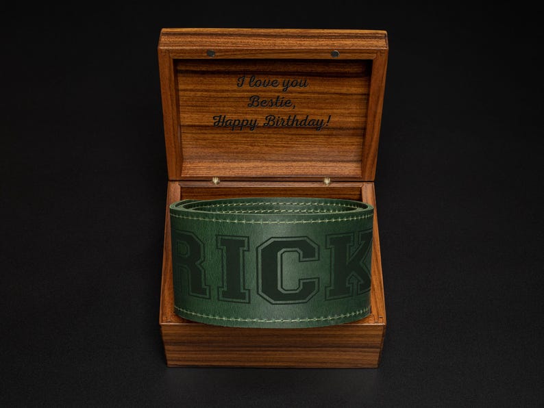 May include: A wooden box with an open lid, revealing a green leather belt with the name "RICK" embossed on it. The inside of the box has an engraved message: "I love you Bestie, Happy Birthday!" The box is made of dark wood.