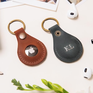 May include: Two leather keychains with metal rings. One keychain is brown with an Apple logo and the other is dark gray with the initials "KL" engraved on it.