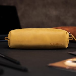 May include: Mustard-colored leather pencil case with the name "SHARON" embossed on the front. The rectangular case has a zipper closure and a small loop on one end. A second, brown leather case is visible in the background.