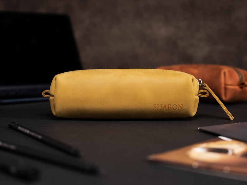 May include: Mustard-colored leather pencil case with the name "SHARON" embossed on the front. The rectangular case has a zipper closure and a small loop on one end. A second, brown leather case is visible in the background.