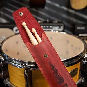 May include: A red leather drumstick case with a flame design, holding two wooden drumsticks. The case is open, revealing the sticks, and is positioned in front of a drum set.