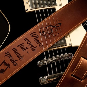 K&ouml;nnte beinhalten: Brauner Leder-Gitarrengurt mit dem Text "Where words fail - music speaks" und Noten. Der Gurt liegt &uuml;ber einer schwarzen E-Gitarre und zeigt die Saiten und die Hardware.
