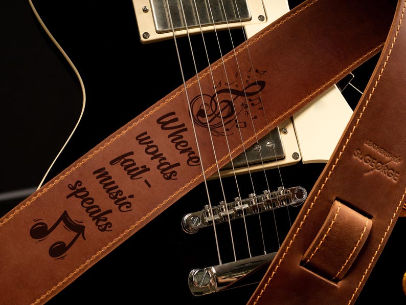 K&ouml;nnte beinhalten: Brauner Leder-Gitarrengurt mit dem Text "Where words fail - music speaks" und Noten. Der Gurt liegt &uuml;ber einer schwarzen E-Gitarre und zeigt die Saiten und die Hardware.