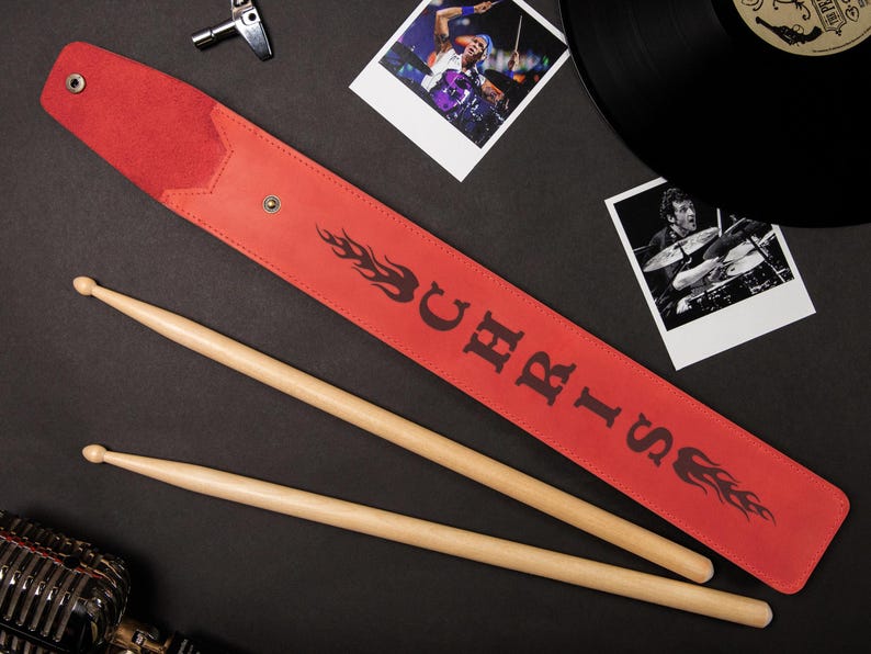 May include: A red leather drumstick holder with the name "CHRIS" in black lettering and flame graphics. Two wooden drumsticks are next to the holder. The background includes a vinyl record and photos.