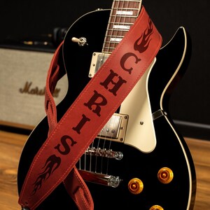 May include: A black electric guitar with a red leather strap. The strap has the name "SIRICH" in large letters and flame designs. The guitar has gold knobs and a cream-colored pickguard. A Marshall amplifier is in the background.