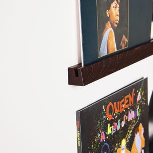 May include: A dark brown wooden record shelf mounted on a white wall, holding two vinyl record album covers. The top album cover features a portrait of a woman in a blue dress. The bottom album cover is black with colorful text and graphics.