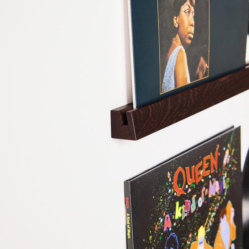 May include: A dark brown wooden record shelf mounted on a white wall, holding two vinyl record album covers. The top album cover features a portrait of a woman in a blue dress. The bottom album cover is black with colorful text and graphics.