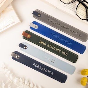 May include: A collection of personalized leather bookmarks in various colors, including gray, blue, and navy. Each bookmark features embossed text with names, dates, or phrases like "Let your mind wander." The bookmarks are arranged near a book and glasses.