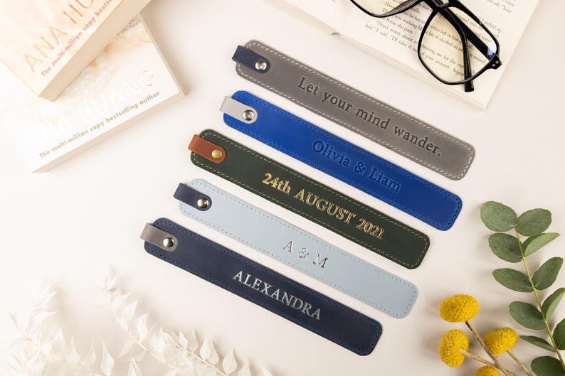 May include: A collection of personalized leather bookmarks in various colors, including gray, blue, and navy. Each bookmark features embossed text with names, dates, or phrases like "Let your mind wander." The bookmarks are arranged near a book and glasses.