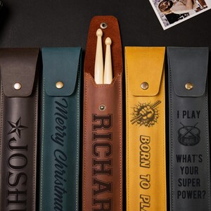 May include: A collection of leather drumstick cases in various colors, including brown, teal, and yellow. Each case features personalized text or graphics, such as a star, the name "RICHARD", and the phrases "Merry Christmas" and "I PLAY".