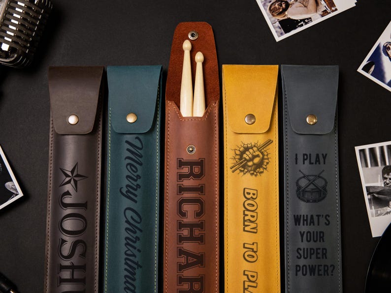 May include: A collection of leather drumstick cases in various colors, including brown, teal, and yellow. Each case features personalized text or graphics, such as a star, the name "RICHARD", and the phrases "Merry Christmas" and "I PLAY".