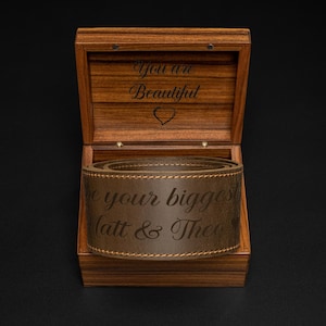 K&ouml;nnte beinhalten: Eine Holzbox mit offenem Deckel, die einen braunen Lederg&uuml;rtel im Inneren zeigt. Die Box ist mit den Worten "You are Beautiful" und einem Herz graviert. Der G&uuml;rtel ist mit "be your biggest Matt & Theo" beschriftet.