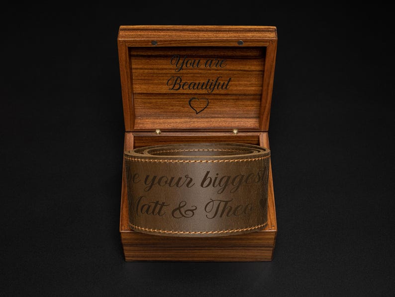 K&ouml;nnte beinhalten: Eine Holzbox mit offenem Deckel, die einen braunen Lederg&uuml;rtel im Inneren zeigt. Die Box ist mit den Worten "You are Beautiful" und einem Herz graviert. Der G&uuml;rtel ist mit "be your biggest Matt & Theo" beschriftet.
