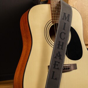 May include: An acoustic guitar with a light wood finish and a black sound hole. A gray guitar strap with the name "MICHAEL" in capital letters is draped across the guitar. The guitar has a brown wood trim.