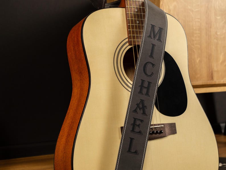 May include: An acoustic guitar with a light wood finish and a black sound hole. A gray guitar strap with the name "MICHAEL" in capital letters is draped across the guitar. The guitar has a brown wood trim.